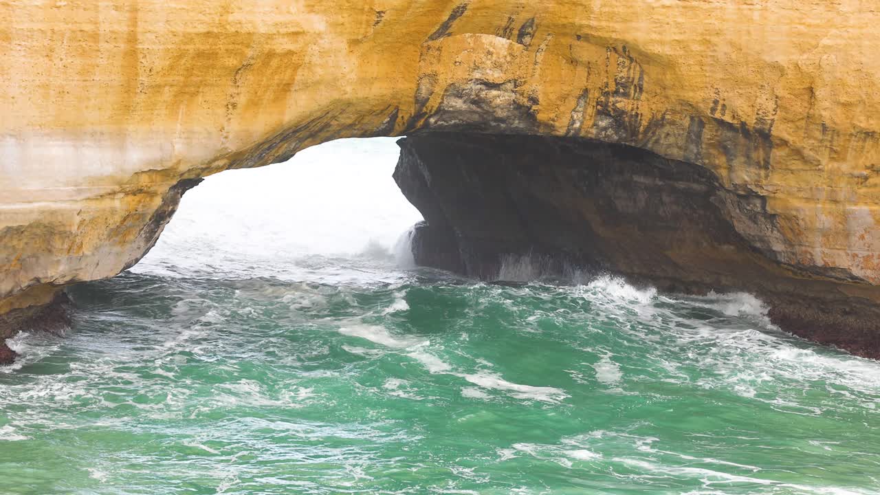 Dynamic ocean waves surge through the natural arch of London Bridge, Port Campbell, under soft daylight, highlighting coastal erosion