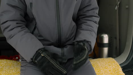 man taking off gloves and making a phone call while sitting in car boot on a snowy winter day