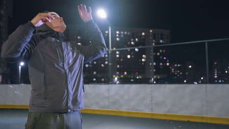 hombre hábilmente lanza la pelota, la equilibra en la espalda, y la empuja hacia arriba, atrapándola con las manos durante la práctica nocturna de fútbol en el campo urbano con luces de la ciudad y fondo iluminado de la calle