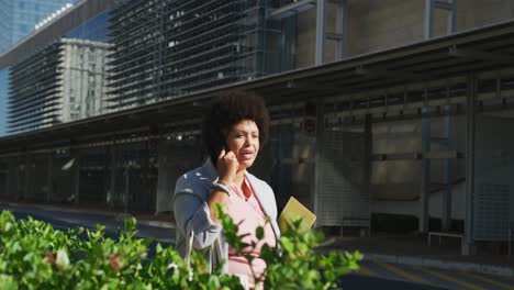 plus size biracial woman talking on smartphone and holding notebooks in city