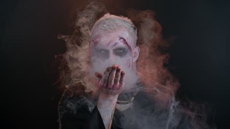 sinister man halloween zombie with bloody face blows smoke from nose and mouth, making air fly kiss