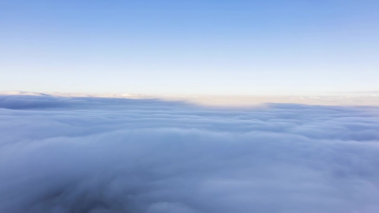 Un movimento fluido in avanti attraversa un'ampia coltre di nuvole pallide mentre un cielo azzurro limpido riempie la parte superiore dell'inquadratura e una luce calda e soffusa traccia l'orizzonte lontano attraverso un paesaggio aperto e calmo