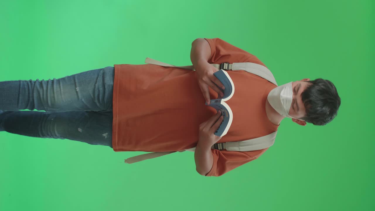 la vista frontal de un estudiante asiático con una máscara, mochila, camiseta marrón y vaqueros azules leyendo un libro mientras camina a la escuela en pantalla verde