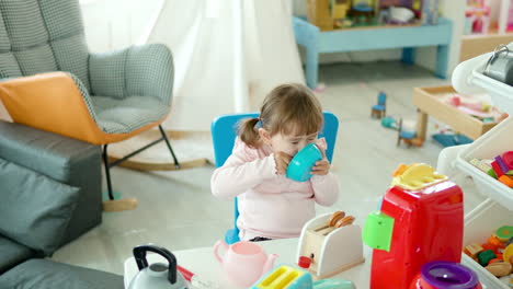 playful little girl pretends eating from toy plate with spoon sitting on chair by the table with kitchenware cooking toys