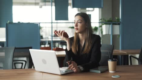 beautiful-young-woman-in-a-stylish-coworking-office-at-her-workspace-records-a-voice-message-on-her-smartphone,-dressed-in-classic-business-attire