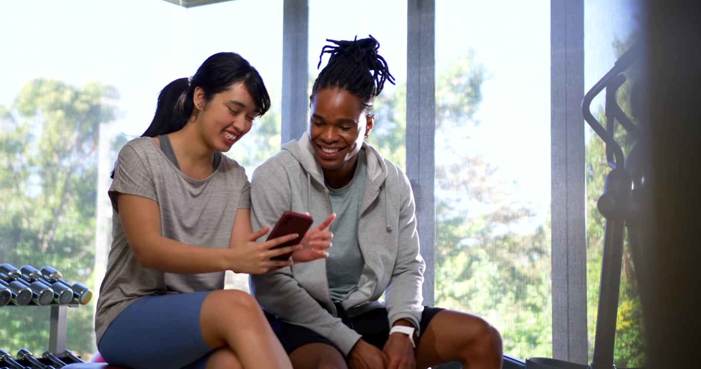 Diverse workout partners in sportswear, woman tilting phone while man leaning, browsing fitness app