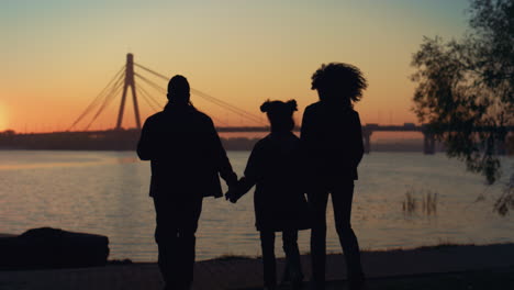 loving family silhouette walking to river shore together at beautiful sunset.