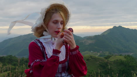 blonde woman dressed in mountain village girl outfit applies lipstick on windy mountain top