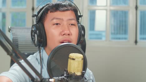 young asian boy in headphones playing the guitar and singing into microphone while recording podcast in studio