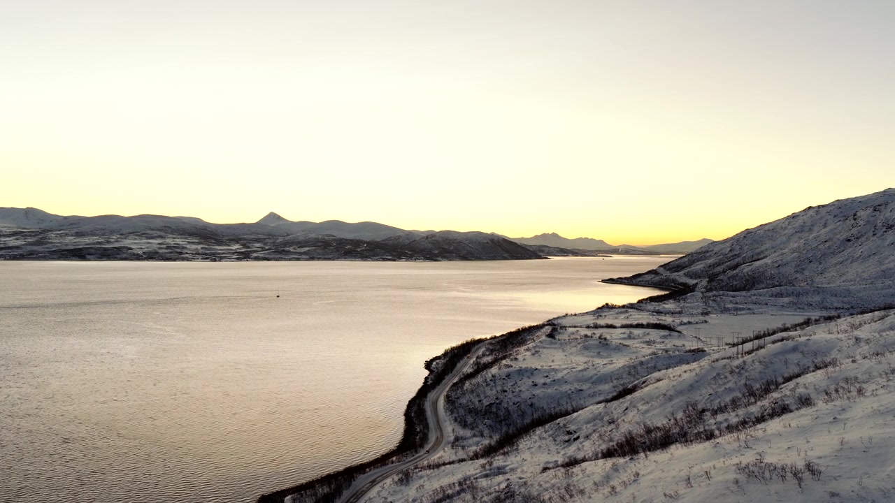 Naturskjønn vei langs fjord i snødekt landskap på Ringvassøya ved solnedgang nær Tromsø, Norge. Luftfoto fremover, plass for tekst.