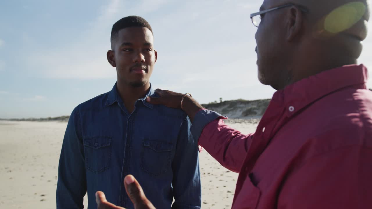 African american father standing on beach with teenage son, putting hand on his shoulder and talking
