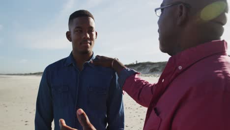African-american-father-standing-on-beach-with-teenage-son,-putting-hand-on-his-shoulder-and-talking