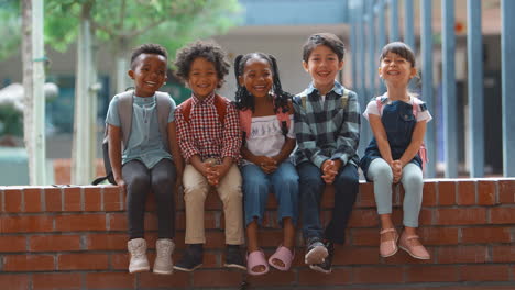 Portrait-Of-Smiling-Multi-Cultural-Elementary-School-Pupils-Sitting-On-Wall-Outdoors-At-School