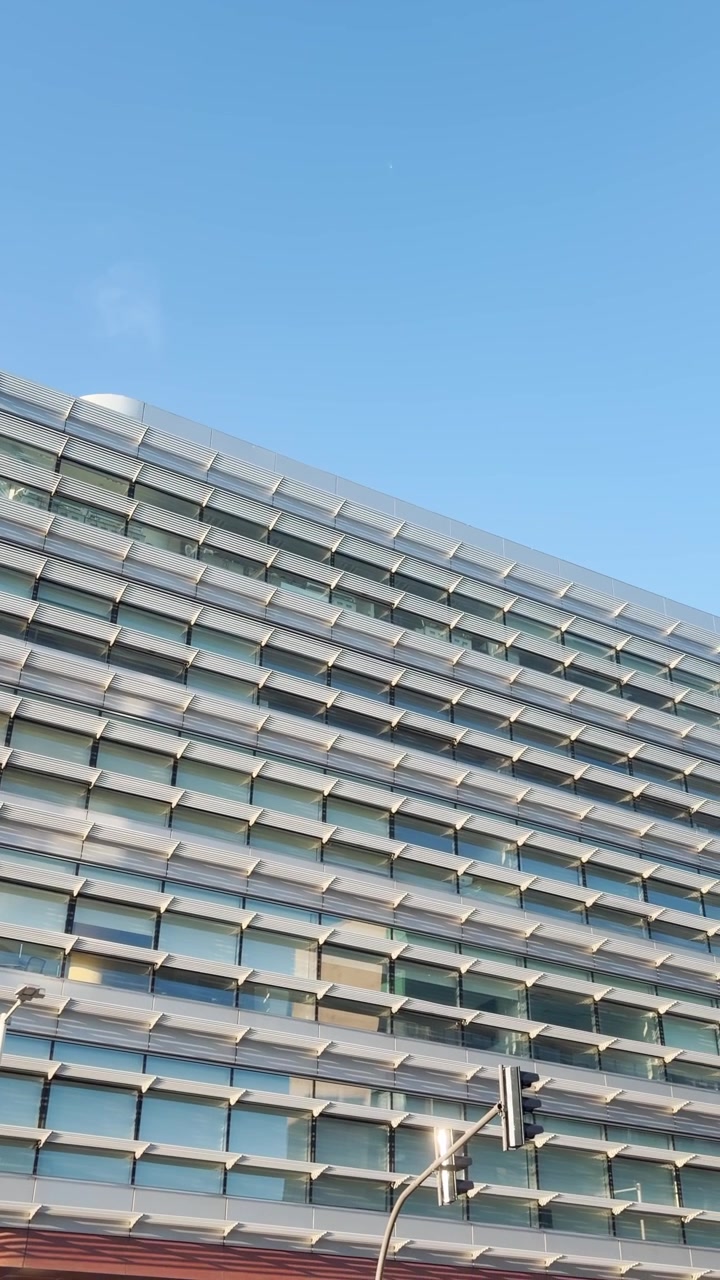 Plano de estabelecimento exterior da fachada de um edifício de escritórios moderno, gravado com câmera de celular em formato de reel vertical para mídia social, estilo UGC. POV de sede ou local de trabalho na cidade em um dia ensolarado ao ar livre.