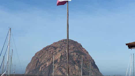 handheld booming down shot of the california state flag with morro rock in the distance in morro bay, california. 4k