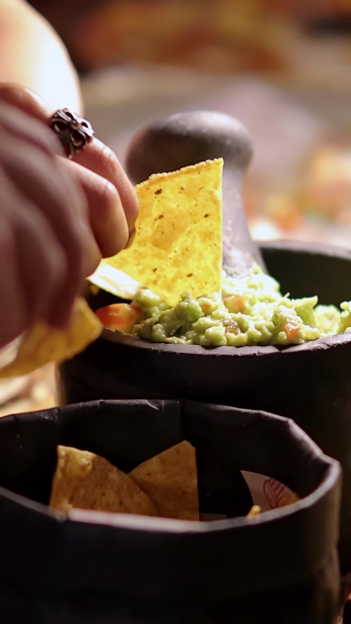 Mani che afferrano tortilla chips e le intingono nel guacamole fresco in un mortaio di pietra, sotto un'illuminazione calda e invitante, suggerendo un raduno festivo.