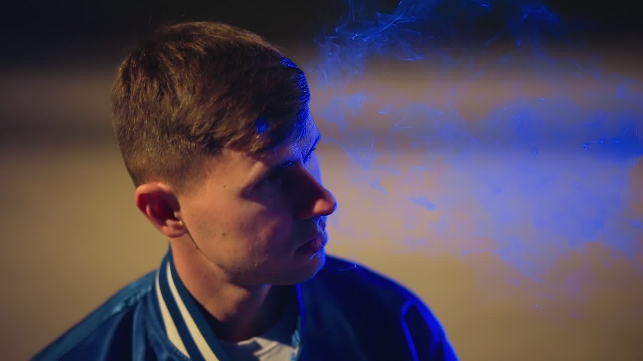 밤 주차장, 흰색 옷을 입은 남자가 전자담배를 피우는 모습, 호박색과 파란색 조명, 손가락은 입 근처, 깊게 들이마시고 천천히 내뱉는 숨, 연기 구름이 흩날리고, 폭격기 재킷, 도시 통근 분위기, 보케 헤드라이트, 반사되는 포장 도로