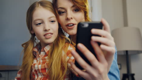 close up of the beautiful daughter and mother sitting close to each other and making selfies on the black smartphone in the room at home. indoors. portrait