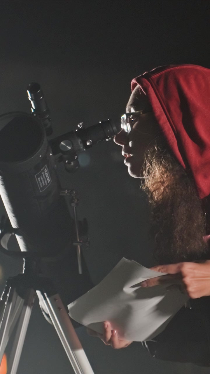 Woman in hooded jacket examines notes near telescope. She wears glasses and has long curly hair looking. Scene includes metal parts from equipment against dark background