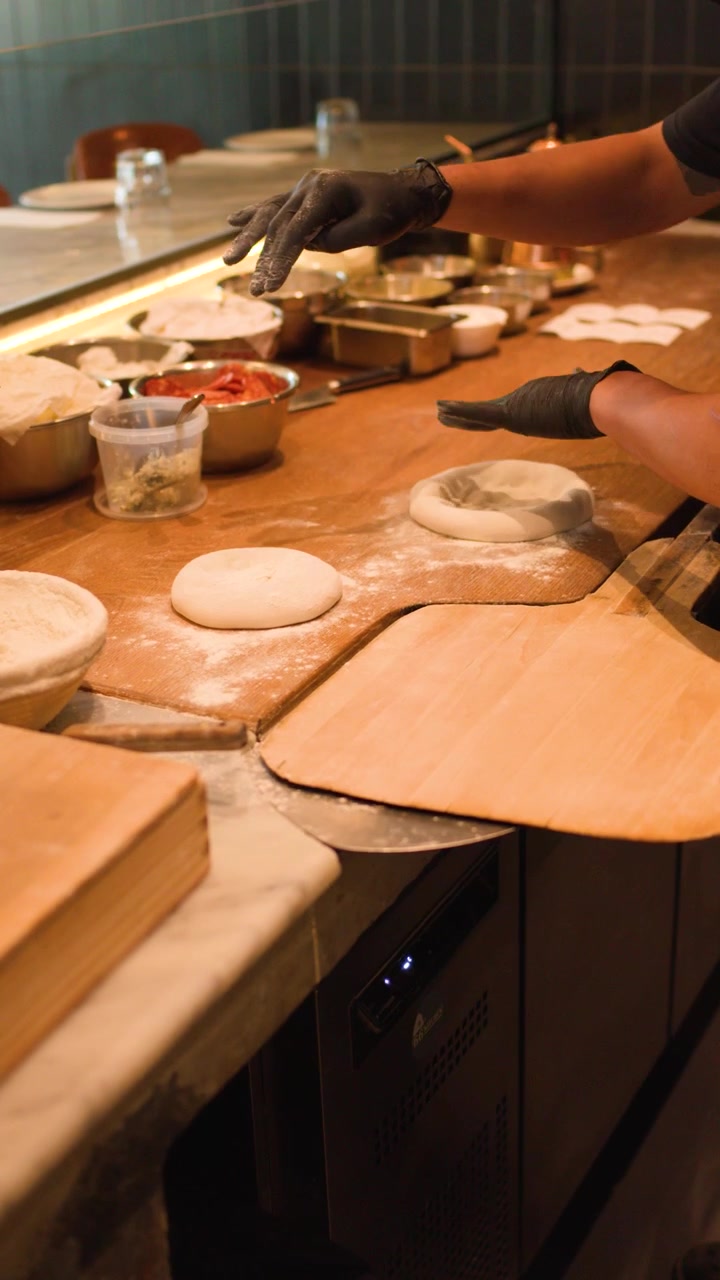 Uno chef che indossa guanti neri prepara l'impasto della pizza su un bancone di legno infarinato a Bangkok.