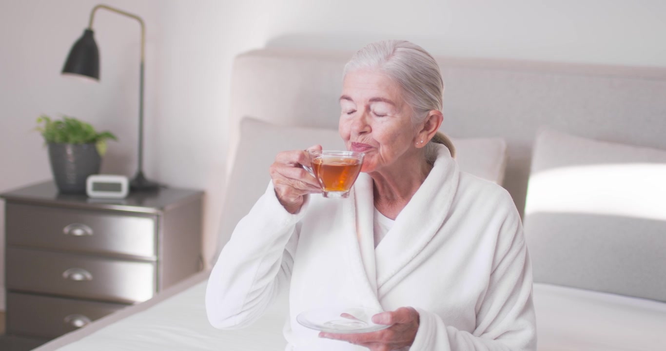 Senhora idosa olhando para a direita, levantando uma xícara de vidro com pires na cama e tomando chá, sorrindo, falando.