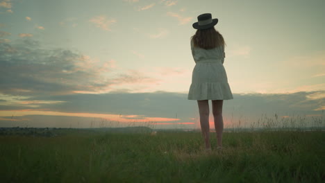 back view of a woman in a white dress and hat, arms folded, standing alone in a serene grassy field, gazing out over a tranquil lake during sunset. as the camera zooms in