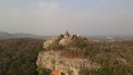 Aerial-dolly-cinematic-shot-of-Wat-Phra-Phutthachai-Buddhist-Temple-located-on-top-of-al-rock-mountain-that-extrude-out-of-dense-tropical-forest,-in-Saraburi,-Thailand