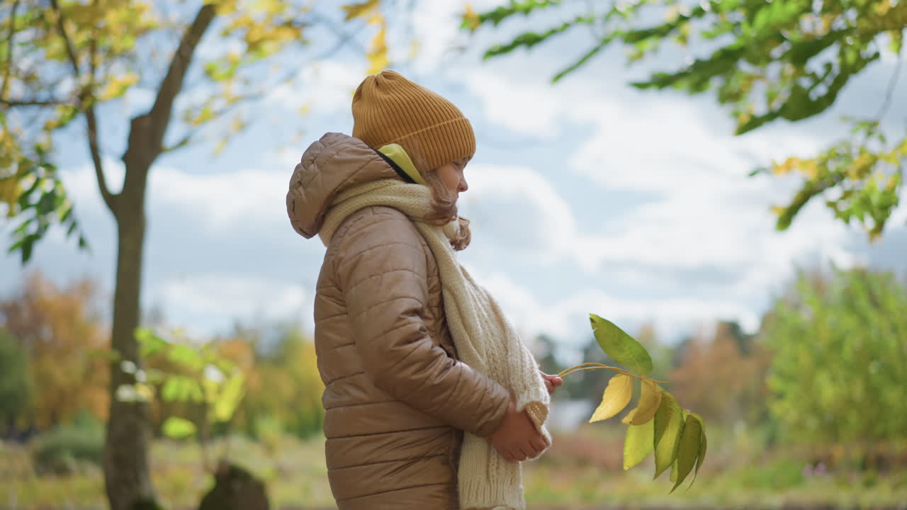side view of cute girl standing holding yellow leaf wearing mustard beanie beige jacket and knitted scarf under tree canopy on leaf strewn park path during crisp fall day with soft cloudy sky