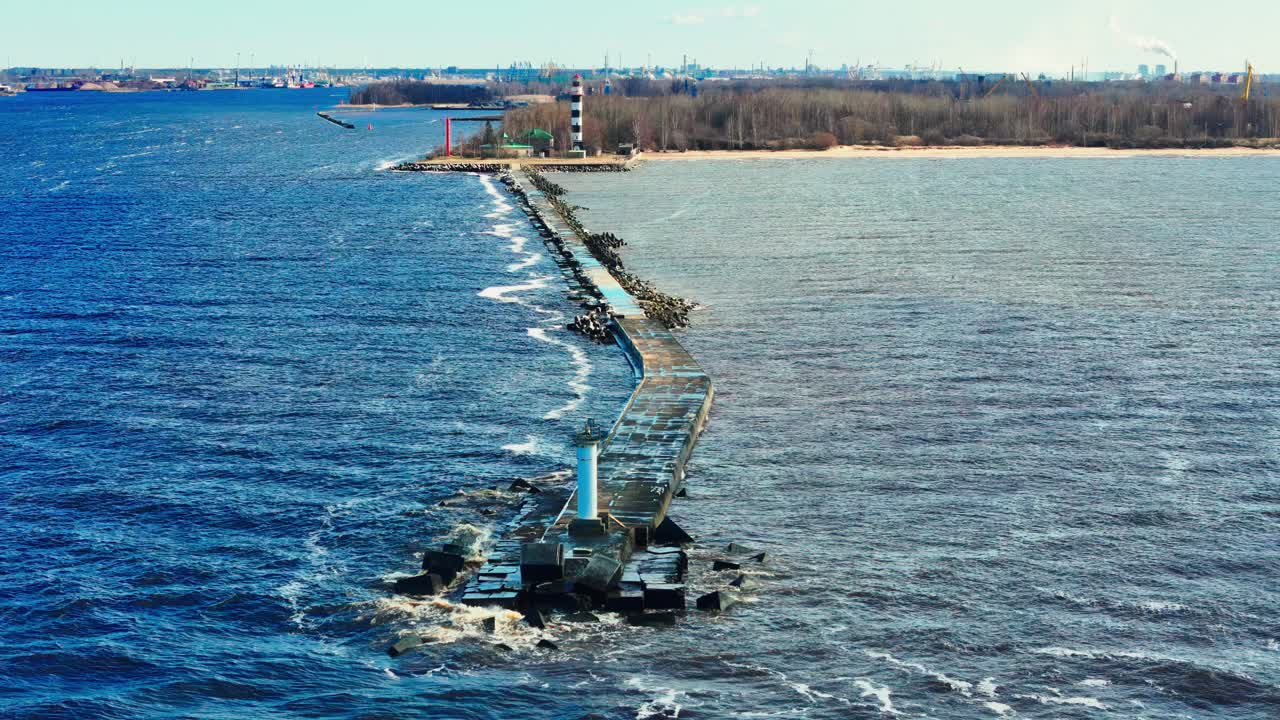 An aerial view of a long concrete pier stretching into the sea, ending at a small white beacon, with a taller striped lighthouse visible inland along a forested shoreline