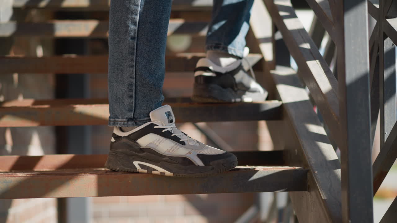 leg view of person wearing blue jeans and white black canvas sneakers descending rusted metal staircase with geometric railing and sunlight casting angular shadows on steps urban outdoor setting