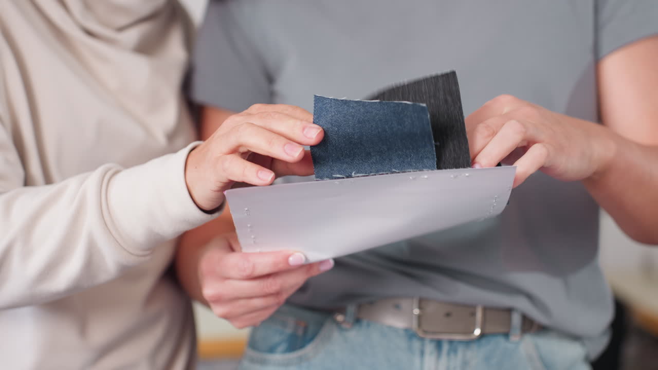 Garment makers standing close together carefully checking different material types from sample sheets during creative collaboration, demonstrating attention to texture, quality, and teamwork