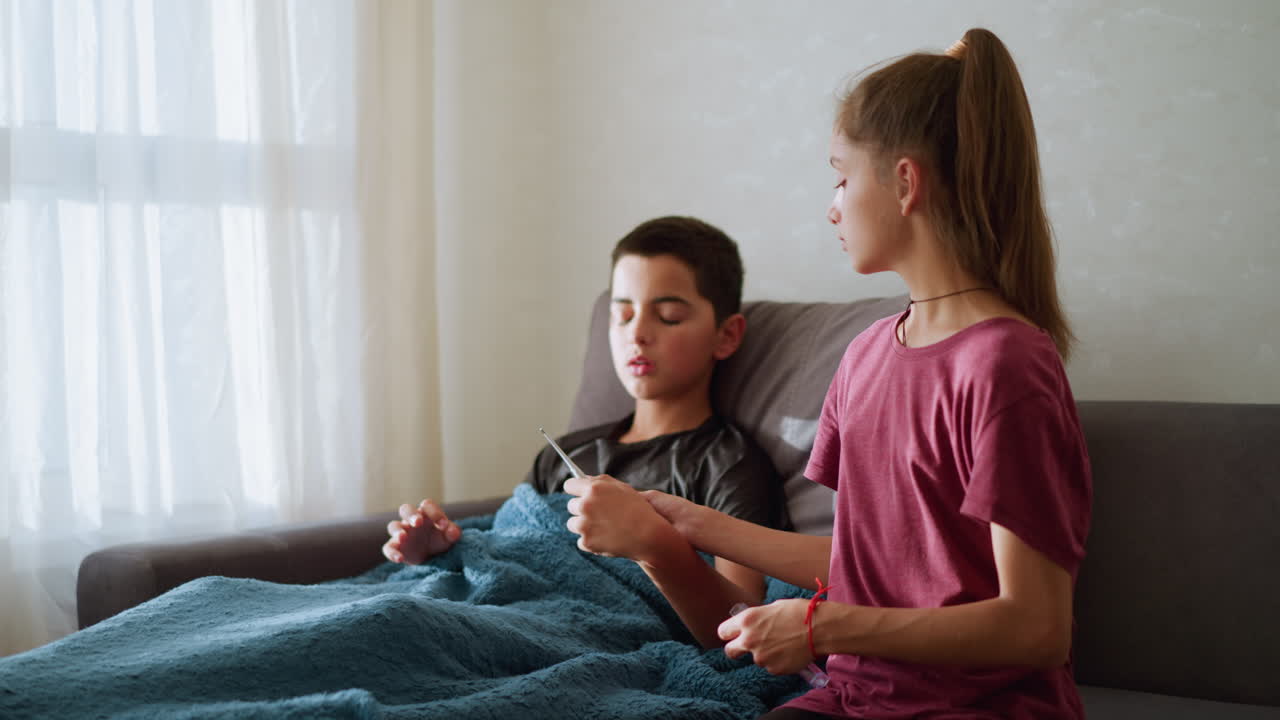 Sick boy sitting on couch collects thermometer from sister wearing rope around neck and wrist, placing it under his armpit to check his temperature, soft natural light coming through window