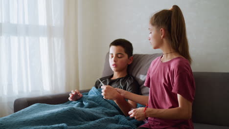 sick boy sitting on couch collects thermometer from sister wearing rope around neck and wrist, placing it under his armpit to check his temperature, soft natural light coming through window