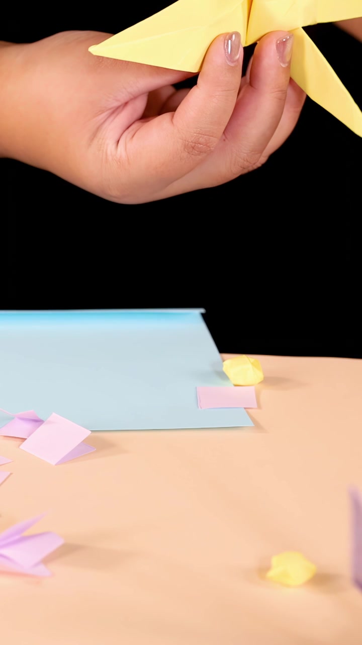 Una persona raccoglie gru di origami e ritagli di carta pastello su un tavolo, sotto un'illuminazione brillante e uniforme.
