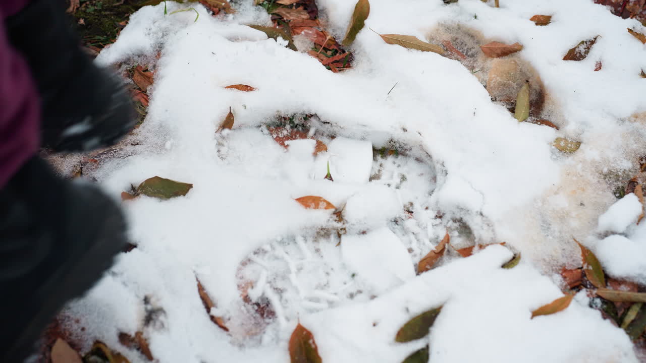 검은색 부츠를 입은 개인이 얼음으로 인 땅 위에 뛰어다니며 어져 있는 건조한 잎과 여러 발자국이 있는 근접 다리 시각