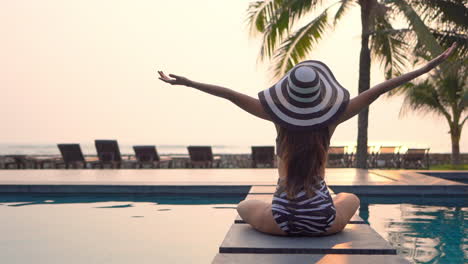 back view of woman with swimsuit and large hat sitting on pool edge and raising arms