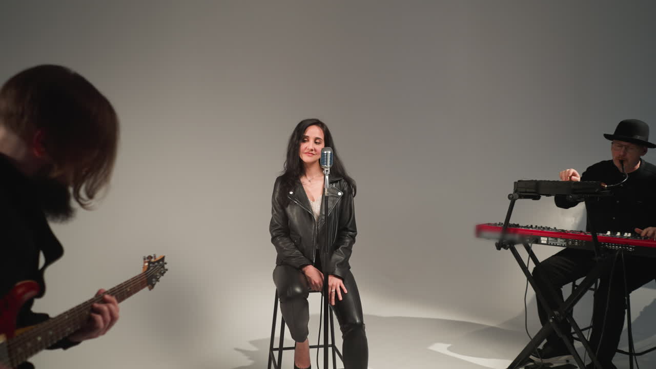 una hermosa vocalista con una chaqueta de cuero negra, sonriendo y cantando con los ojos cerrados, de pie en un micrófono vintage en un taburete alto. un guitarrista toca una guitarra roja y un tecladista trabaja un sampler rojo