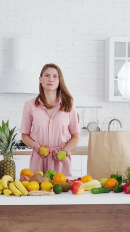 smiling woman with fresh apples. happy housewife standing near the table full of organic fruit and vegetables in the modern kitchen. concept of healthy food. vertical video