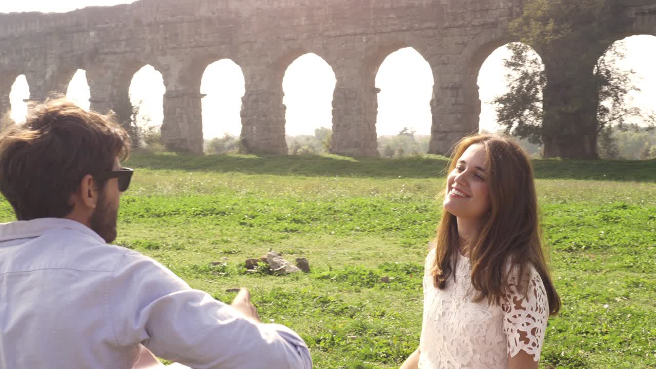 행복한 젊은 사랑스러운 부부는 잔디에 담요 위에 누워 앉아 로마의 고대 아쿠에듀크 앞에 있습니다. 로마의 파르코 데글리 아쿠에두티 공원의 폐허. 로맨틱한 웃음. 기타를 연주하고 노래합니다. 아름다운 소녀. 모자를 던집니다.