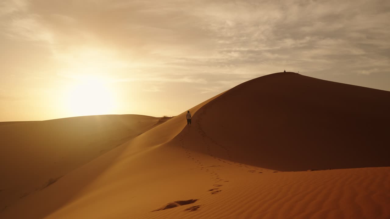 A solitary traveler ascends a golden sand dune in the Sahara Desert during sunrise, leaving a winding trail of footprints under the warm Moroccan sky