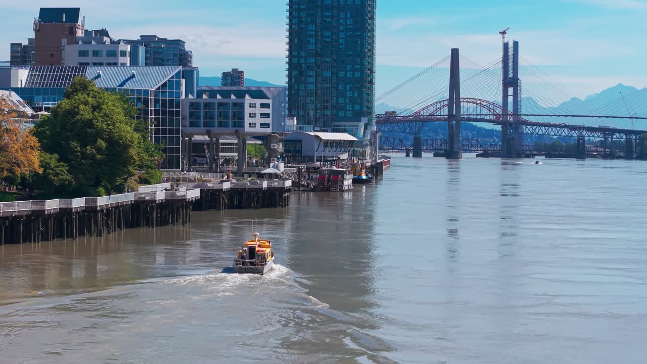 Yüksek binaların ve arka planda Pattullo Köprüsü'nün bulunduğu, British Columbia, New Westminster şehir merkezinin silüetinin önünden geçen Fraser Nehri boyunca ilerleyen bir su taksinin drone çekim görüntüsü