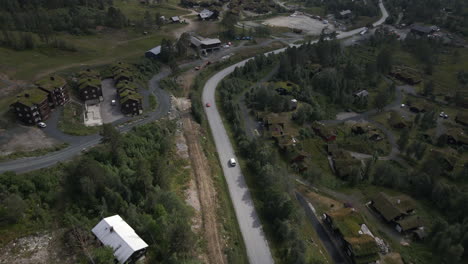 drone following a car which is driving through the road which is surrounded by trees and few residential houses can be seen on the side