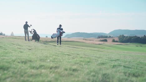 Two-golfers-walking-on-golf-course-carrying-clubs,-big-landscape-overlooking-field