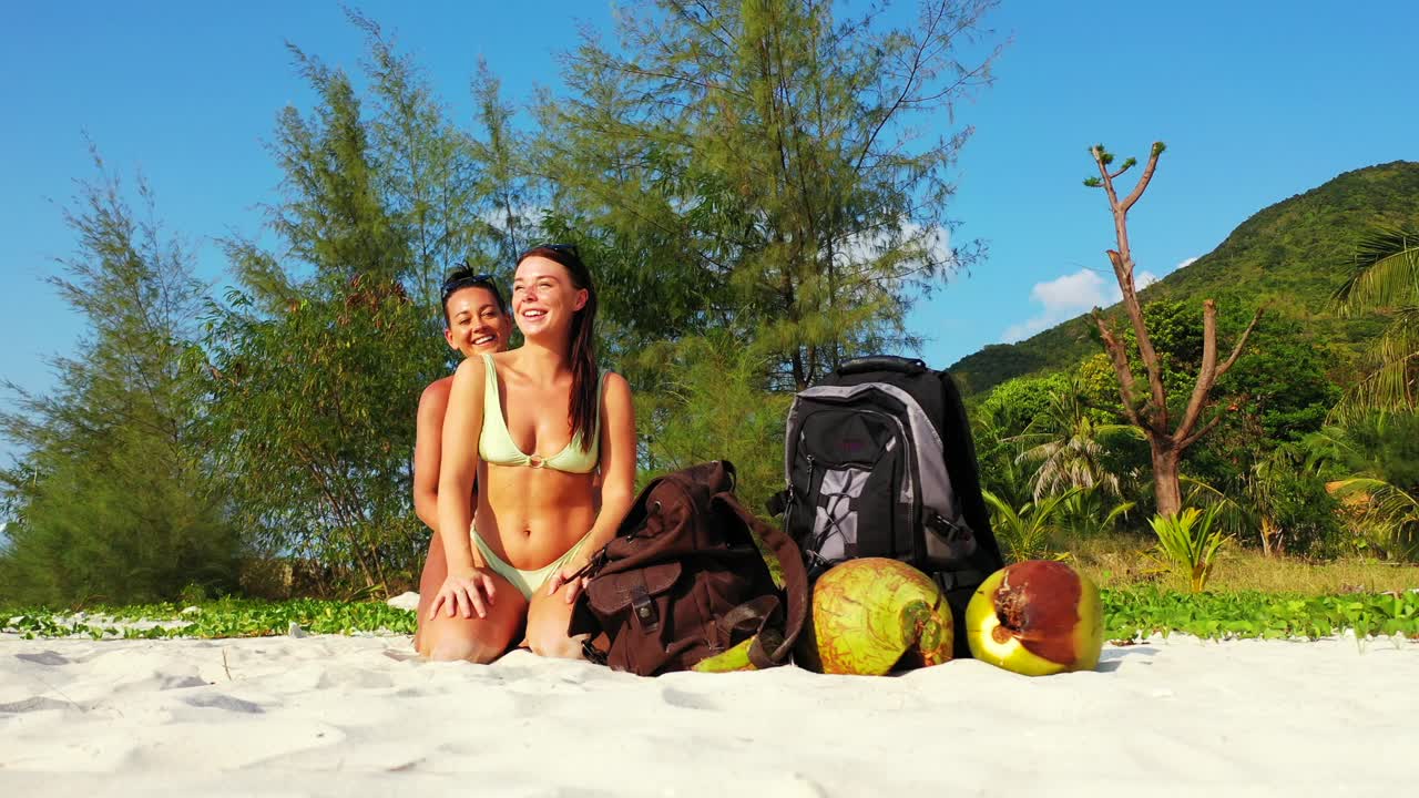 Giovani donne felici in bikini che si massaggiano su una spiaggia esotica vicino a borse da viaggio e noci di cocco lasciate sulla sabbia bianca con sfondo di vegetazione tropicale alle Seychelles