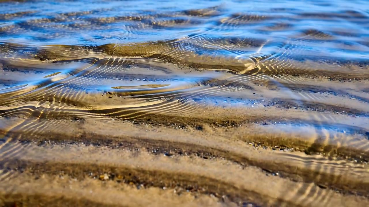 Arriving small wavelets moving shallow seawater over rippled sand ridges on beach, forming caustics