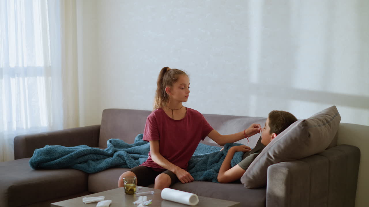 Caring sister checks thermometer used by sick brother resting under blue blanket on couch during quiet indoor moment filled with warmth empathy love and family support in natural daylight environment