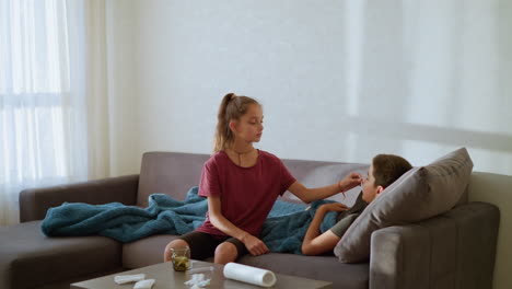 caring sister checks thermometer used by sick brother resting under blue blanket on couch during quiet indoor moment filled with warmth empathy love and family support in natural daylight environment