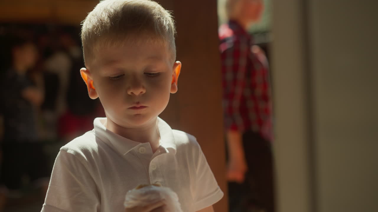 금발 소년은 시장에서 관광객들이 몰려있는 흐릿한 배경에서 식욕이있는 반죽으로 소시지를 먹고 있습니다. 아이는 짜증나는 분위기에서 재미있는 얼굴을 만니다.