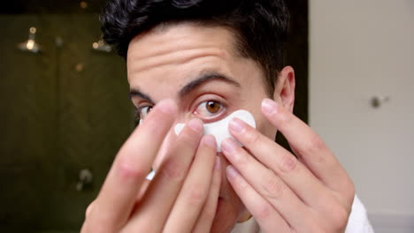 Focused-biracial-man-applying-under-eye-mask-looking-in-bathroom-mirror,-slow-motion