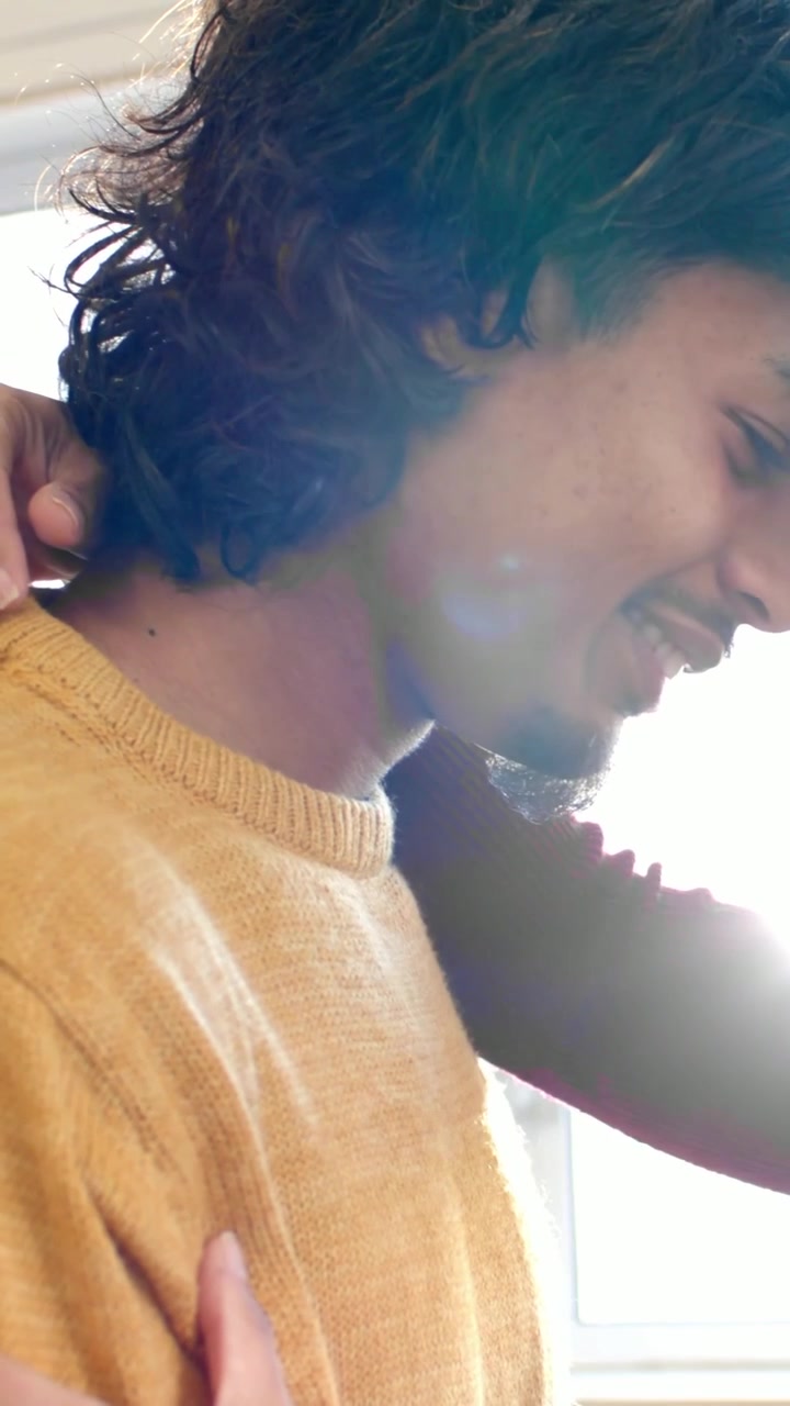 Vertical video: Embracing partner wrapping arms around man by window in mustard sweater, lens flare